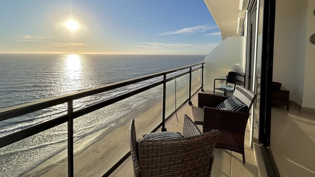 A balcony with wicker furniture overlooking a beach at sunset.