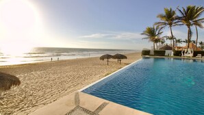 A pool adjacent to a beach with palm trees and a thatched-roof structure.