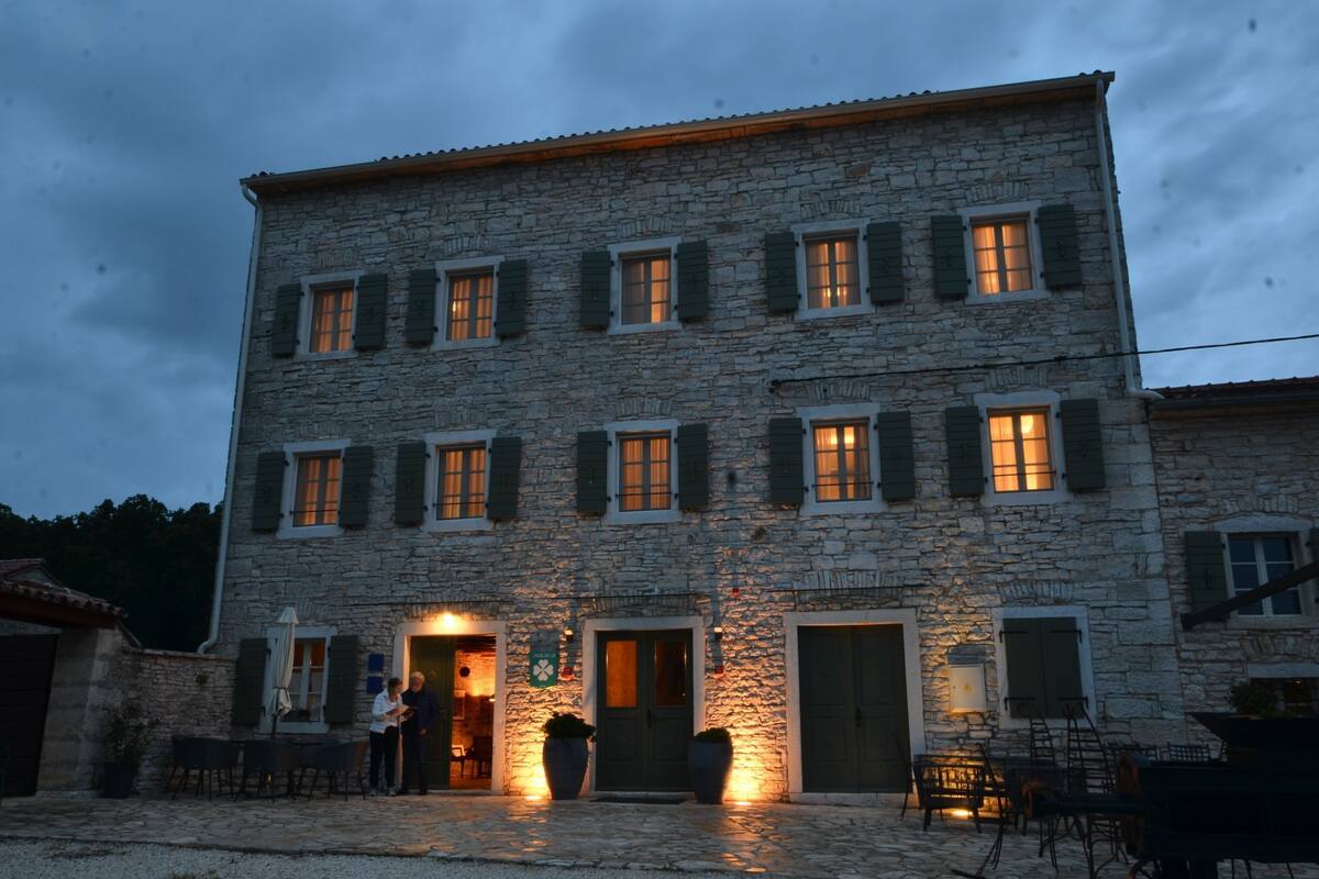 Un bâtiment en pierre doté de nombreuses fenêtres et portes, avec une cour en gravier et des espaces de repos extérieurs.
