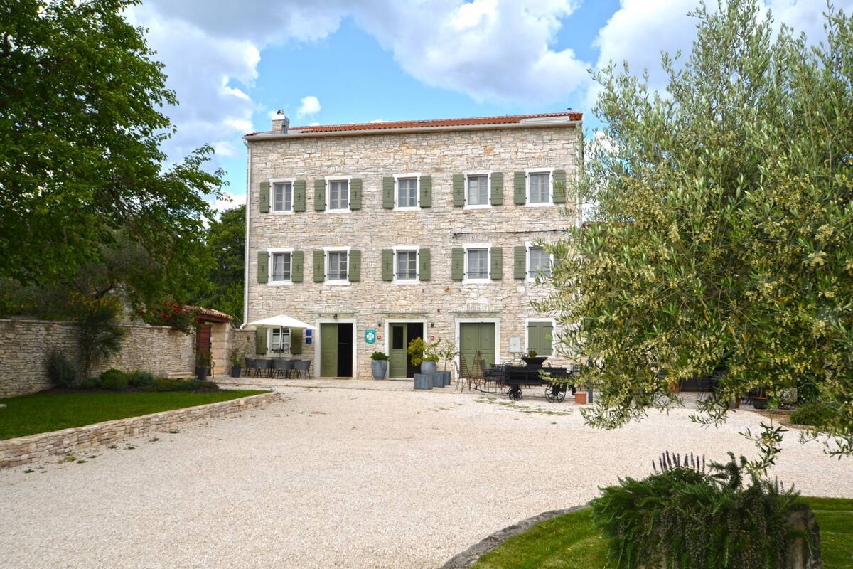 Un bâtiment en pierre doté de nombreuses fenêtres et portes, une cour en gravier et des espaces de repos en plein air.
