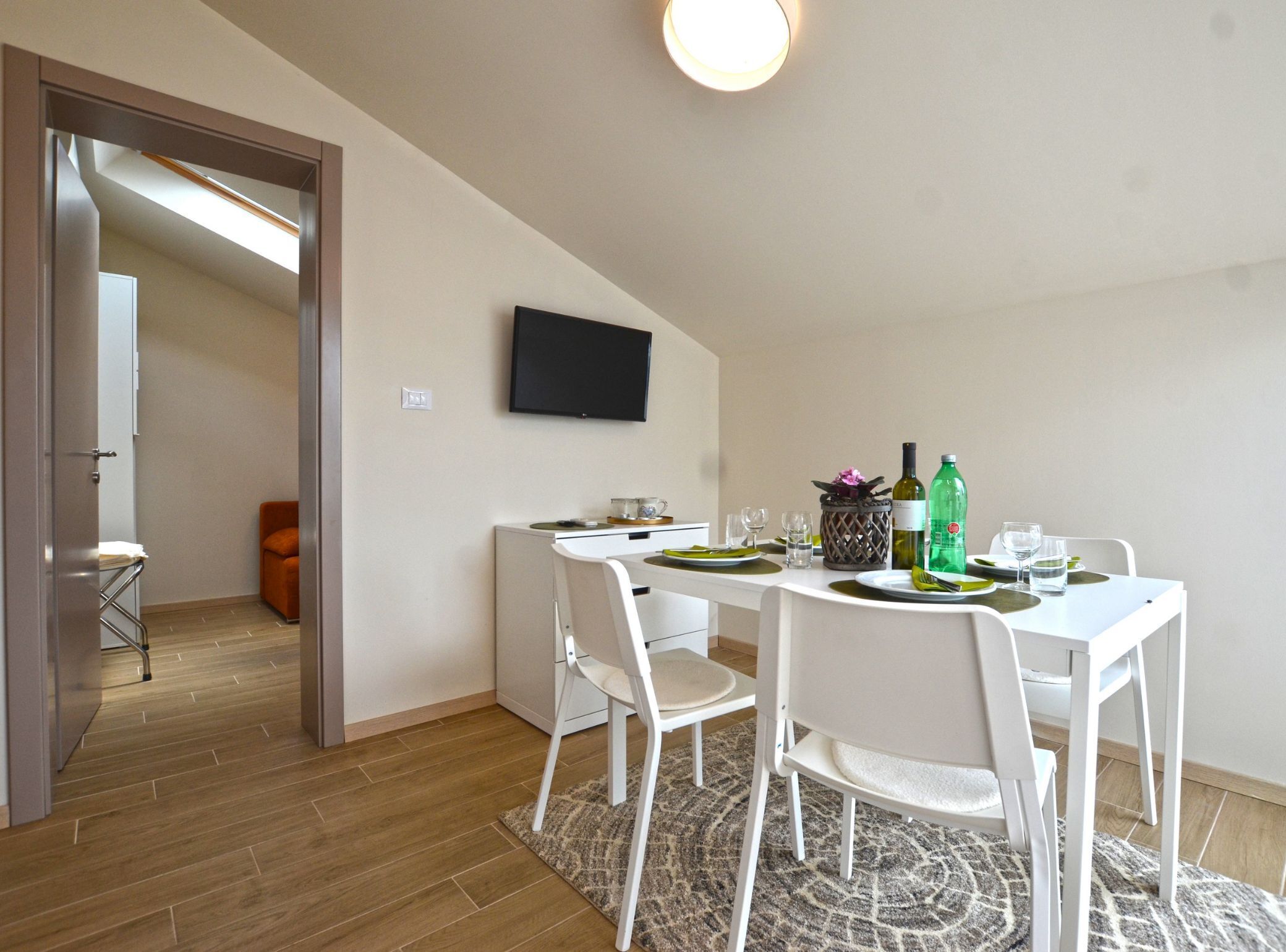 A modern dining area with a white table set for four, a flat-screen TV mounted on the wall, and a wooden floor.