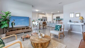 A modern living room with a large TV, comfortable seating, a wooden coffee table, and a kitchen in the background.