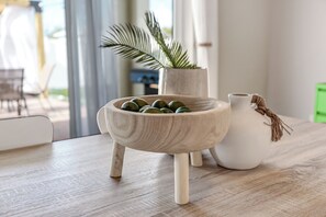 Un cuenco de madera con frutas verdes sobre una mesa de madera, un jarrón blanco con una cuerda, y una planta en maceta al fondo.