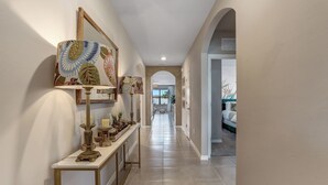 A hallway with a decorative console table, wall-mounted artwork, and a view into a room with a bed and a window.