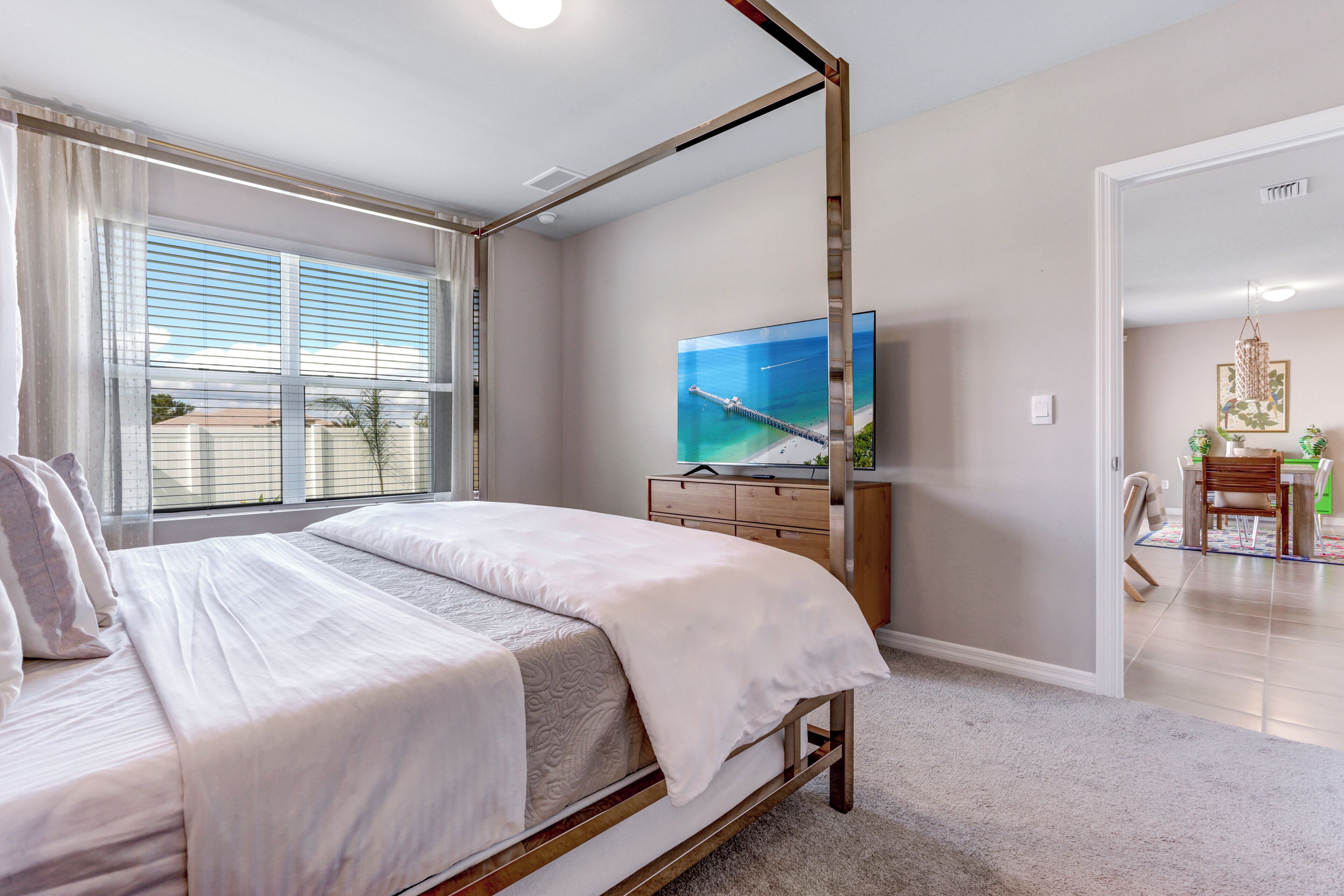 A bedroom with a four-poster bed, a television on a wooden dresser, and a view of the outdoors through large windows.