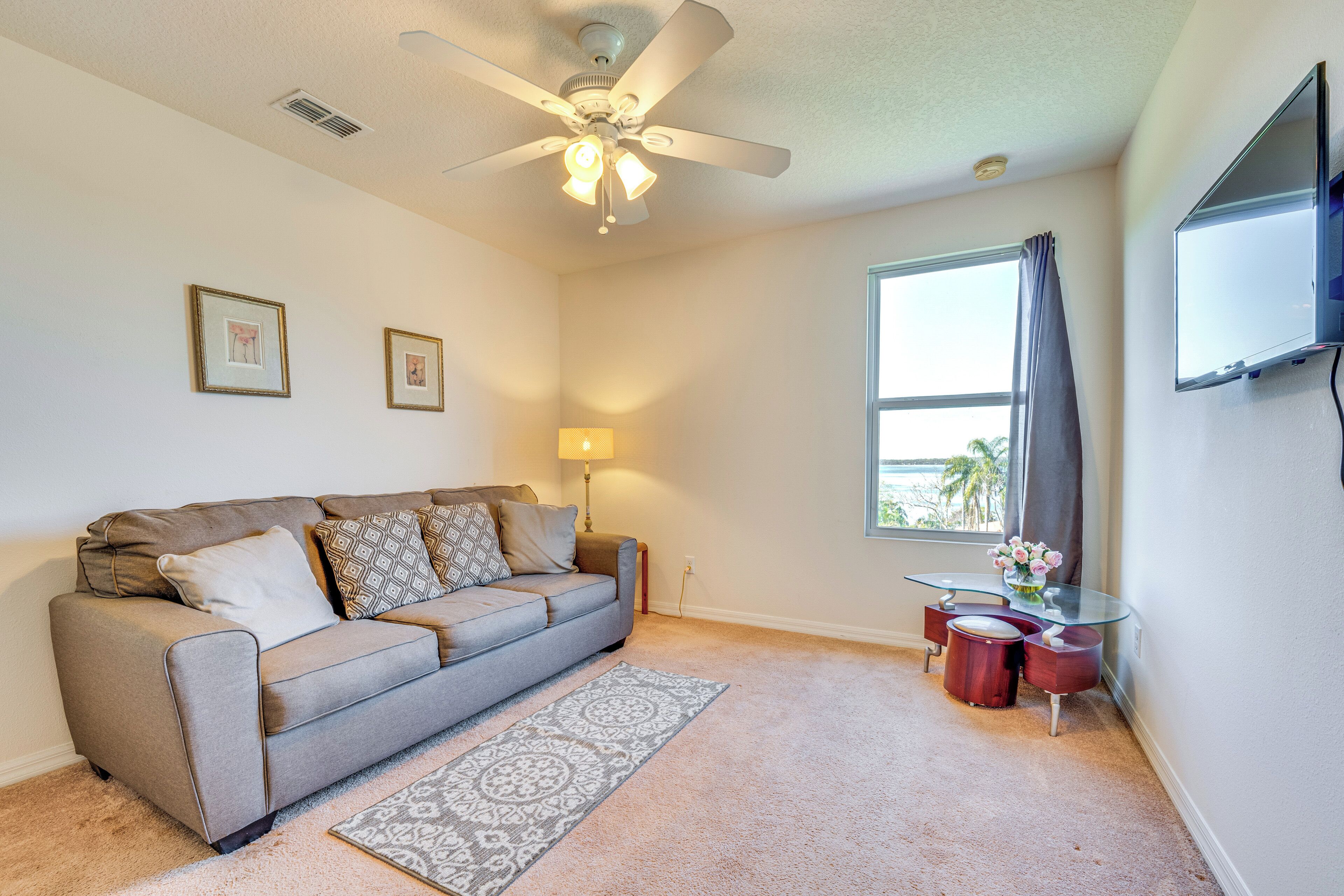 A living room with a grey sofa, a ceiling fan, a lamp, a glass table with a vase of flowers, and a large window with a view of palm trees.