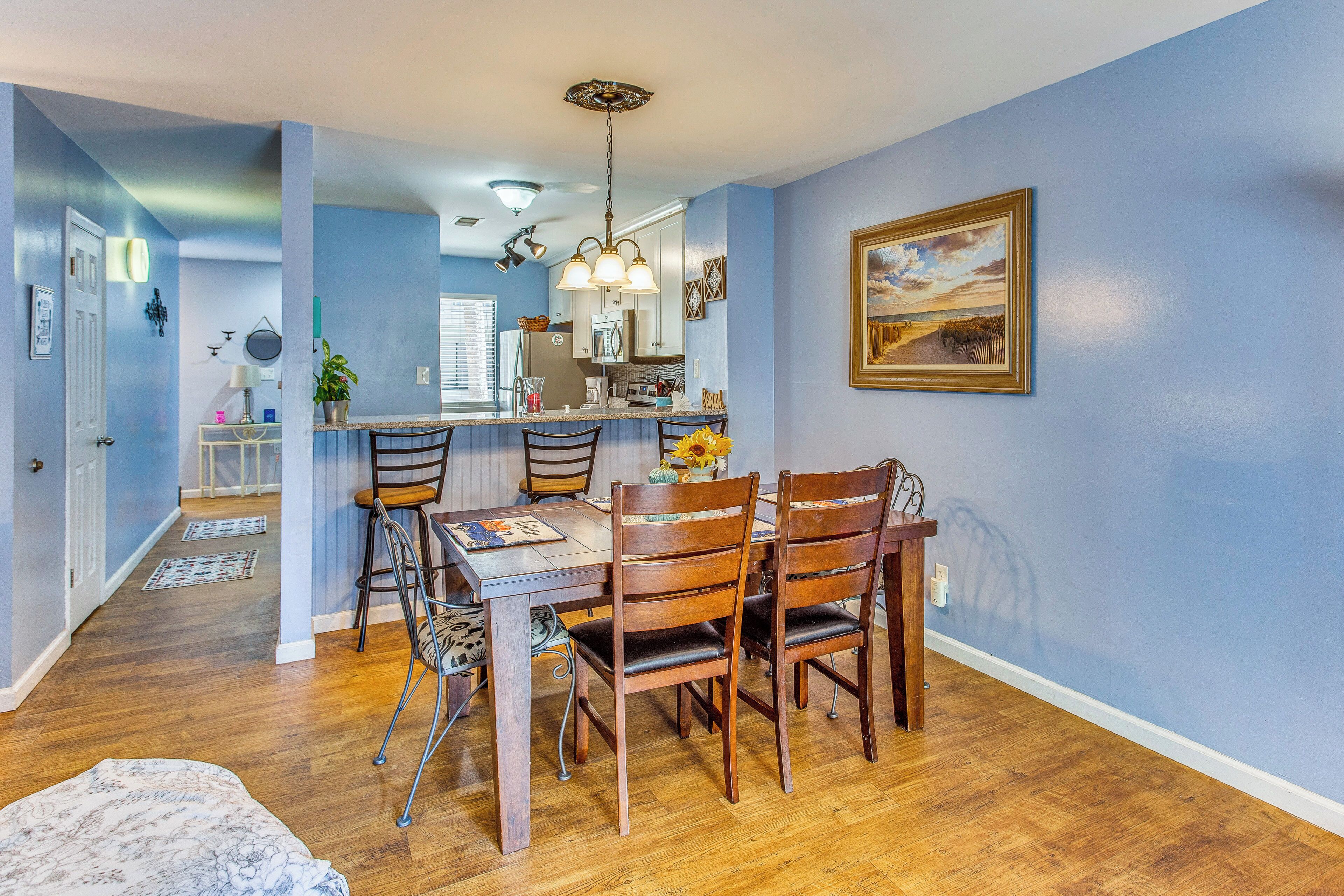 Un comedor con una mesa y sillas de madera, una cocina al fondo y una pared azul con un cuadro enmarcado.