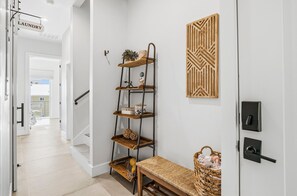 A hallway with a laundry sign, a multi-tiered metal shelf with decorative items, a woven bench, and a patterned wall decoration.