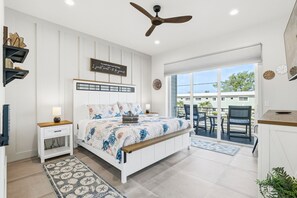 Ein Schlafzimmer mit Bett, Nachttischen, Deckenventilator, großem Fenster und einem Balkon mit Gartenmöbeln.