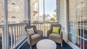 A balcony with wicker chairs and a round table, overlooking a street with buildings and trees.