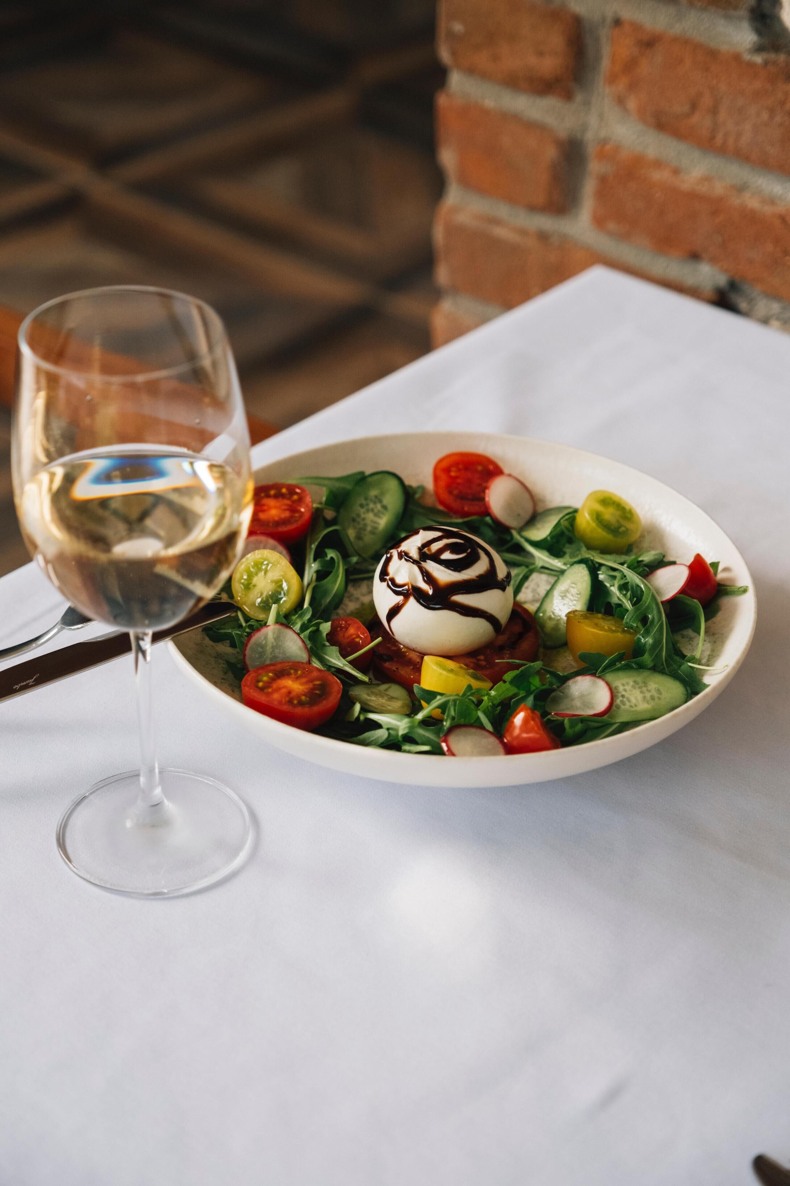 Een witte schaal met salade, een glas witte wijn, en een bakstenen muur op de achtergrond.