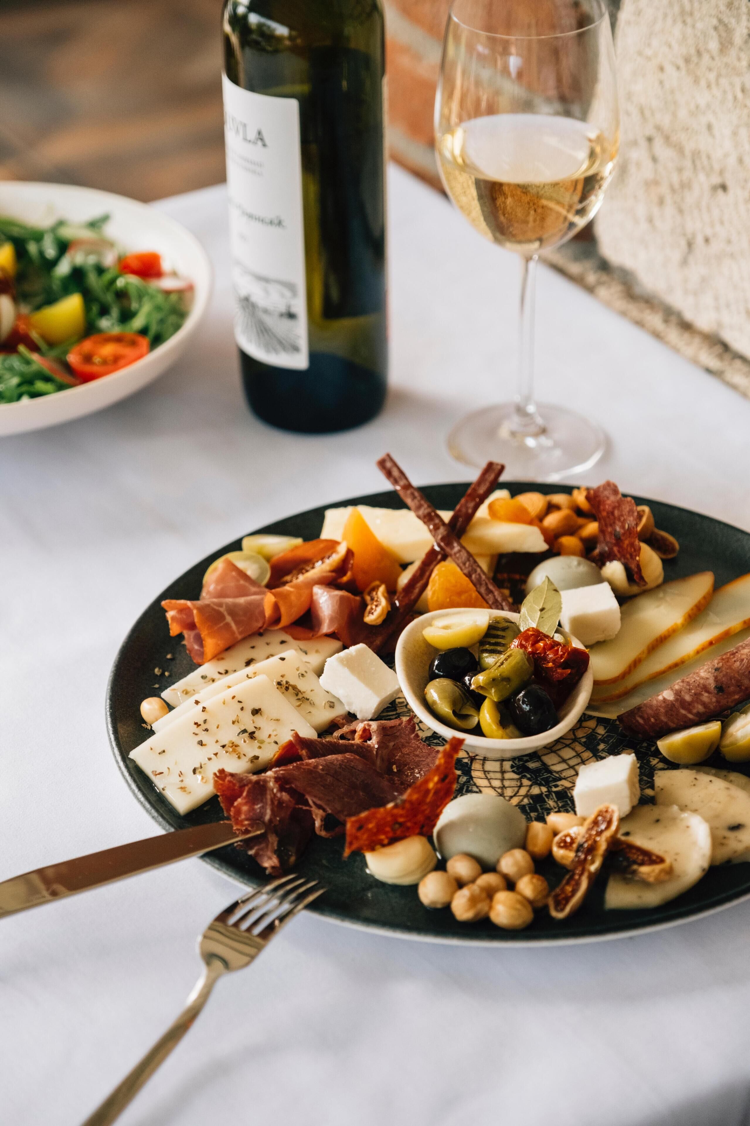 Una tabla de embutidos con varios quesos, carnes y aceitunas en un plato, acompañada de una botella de vino y una copa de vino.