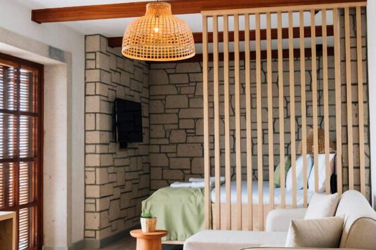 A modern bedroom with a stone accent wall, a wooden slat partition, a green bed, a beige sofa, and a woven rug.