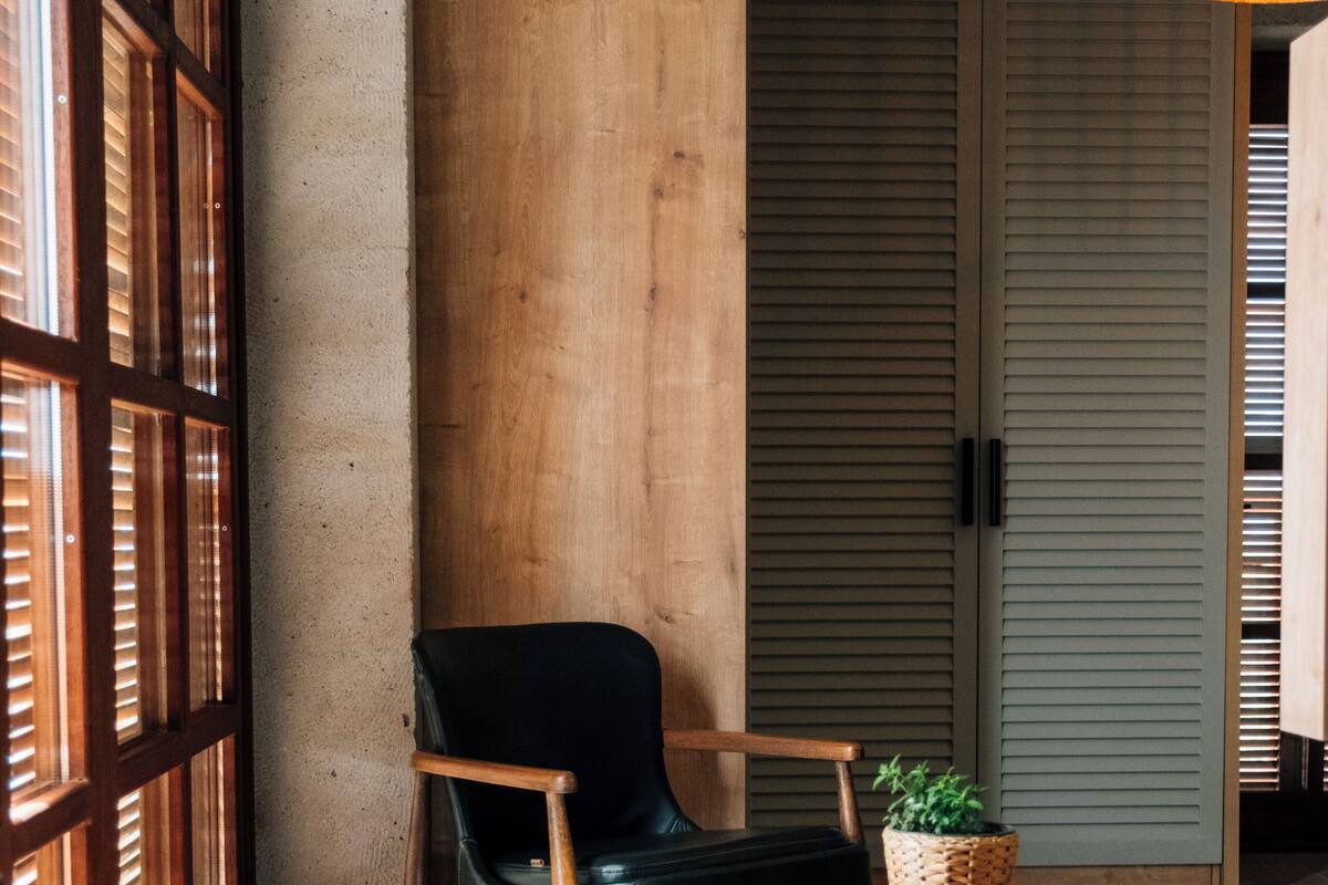 A room with a wooden floor, a black chair, a round wooden table with a plant, and a woven rug.