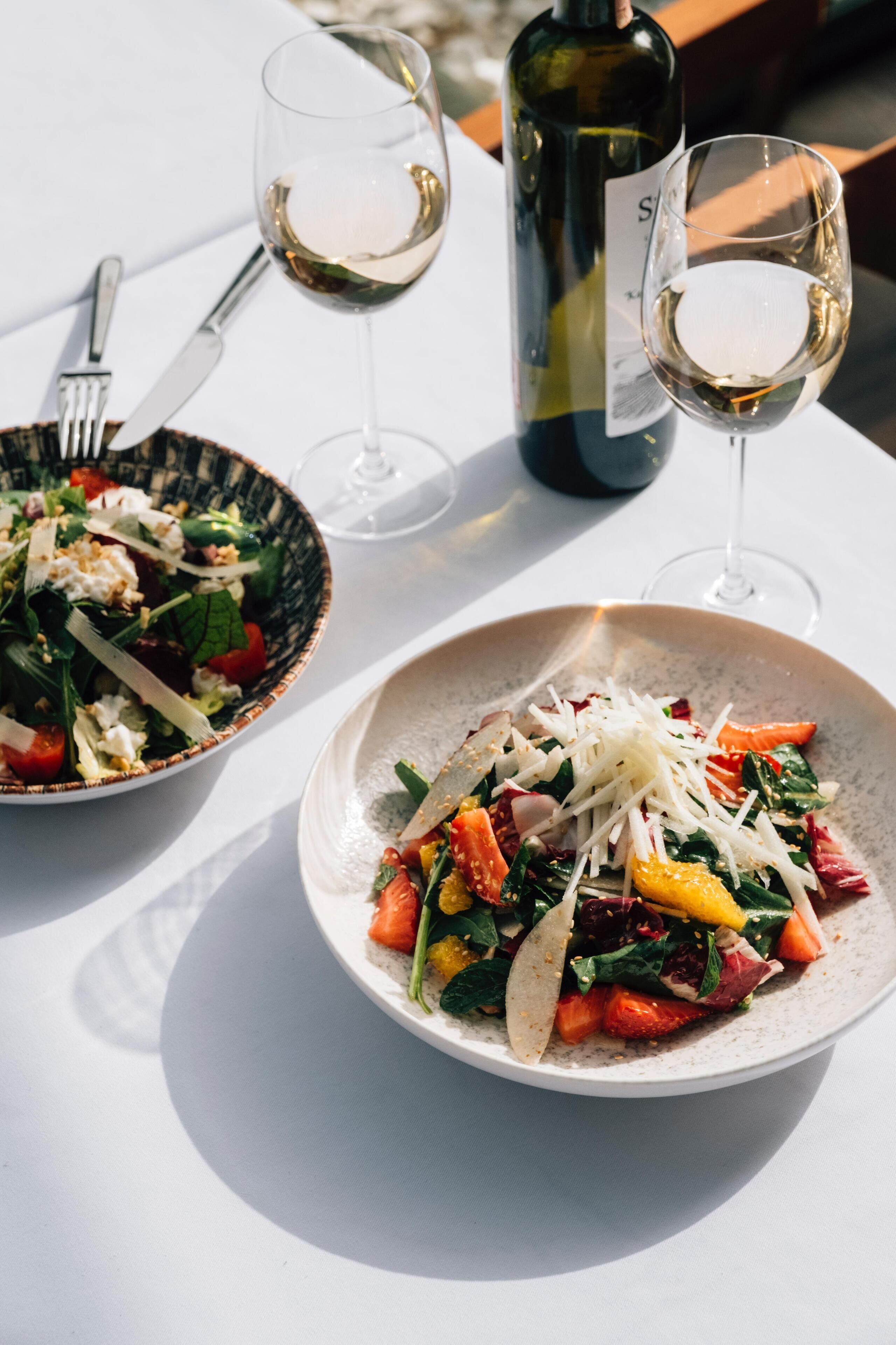 Two plates of salad with a bottle of wine and glasses on a table.