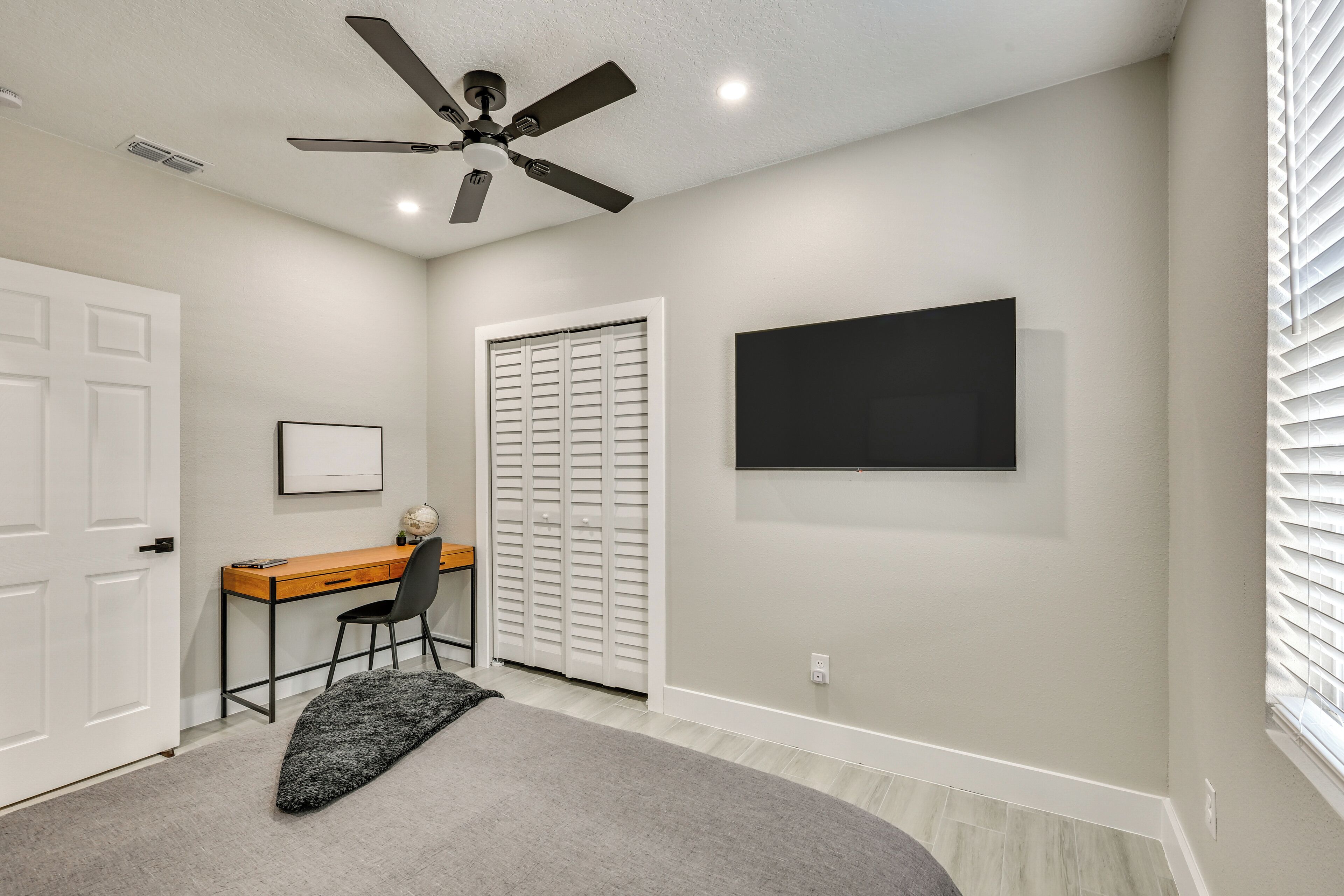 A room with a ceiling fan, a desk with a chair, a closed closet, and a mounted TV.