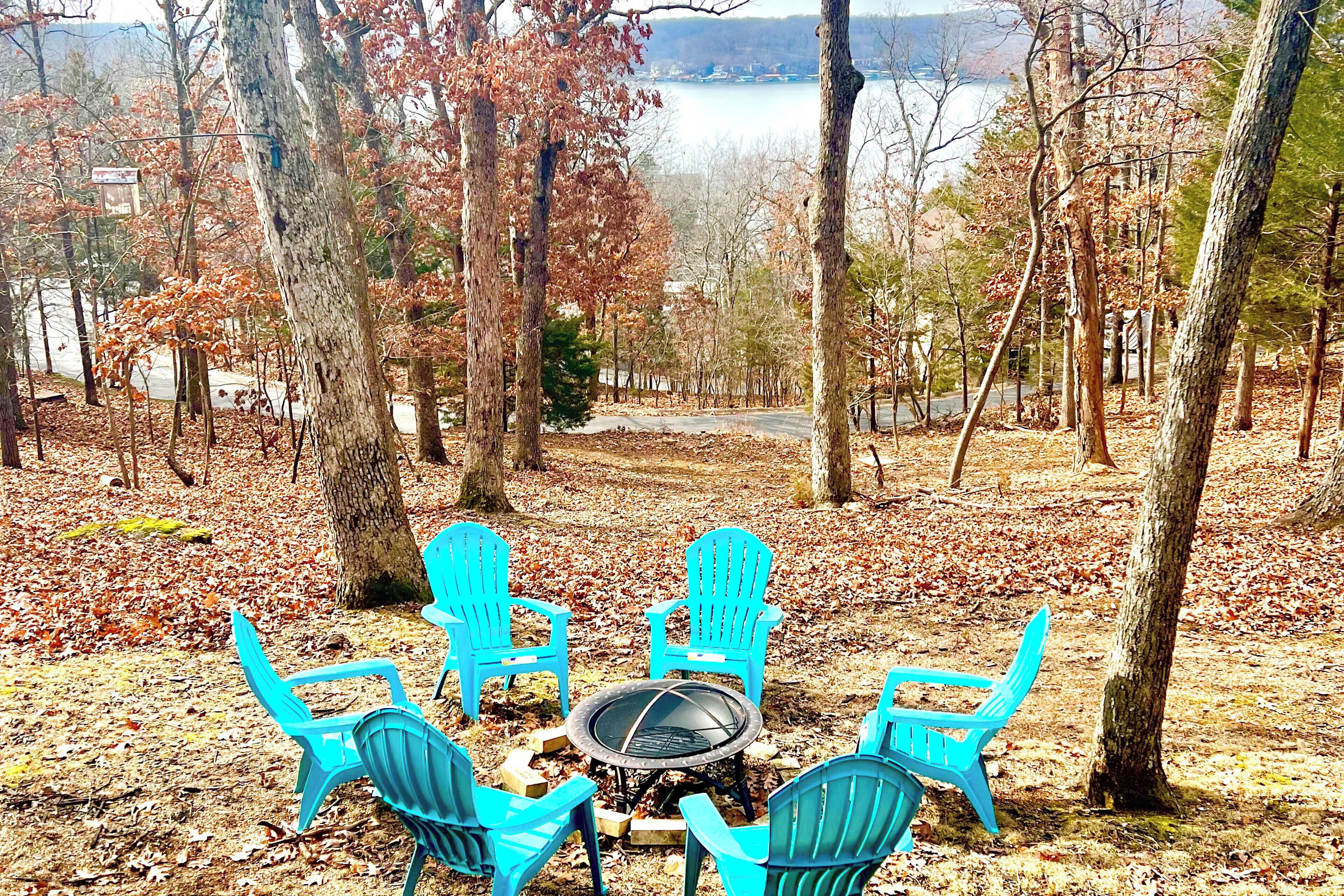 A campsite with four blue Adirondack chairs around a central fire pit, surrounded by trees and a lake view.