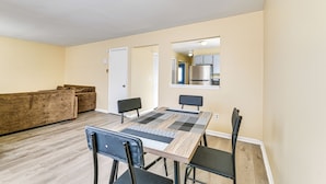 Uma sala de estar com sofá, uma mesa de jantar com cadeiras e uma cozinha visível por uma porta aberta.