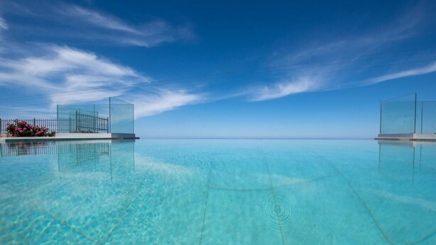 Ein klarer Infinity-Pool mit Blick auf den Ozean und eine gläserne Struktur mit rosa Blüten.