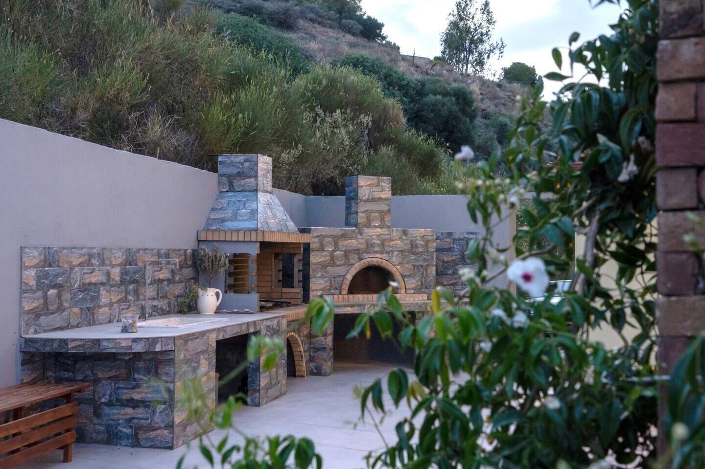 Uma área de cozinha externa com lareira de pedra, forno a pizza e uma mesa de madeira.