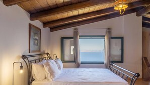 Ein Schlafzimmer mit Bett, Nachttisch, Lampe, einem Fenster mit Vorhängen und Blick aufs Meer.