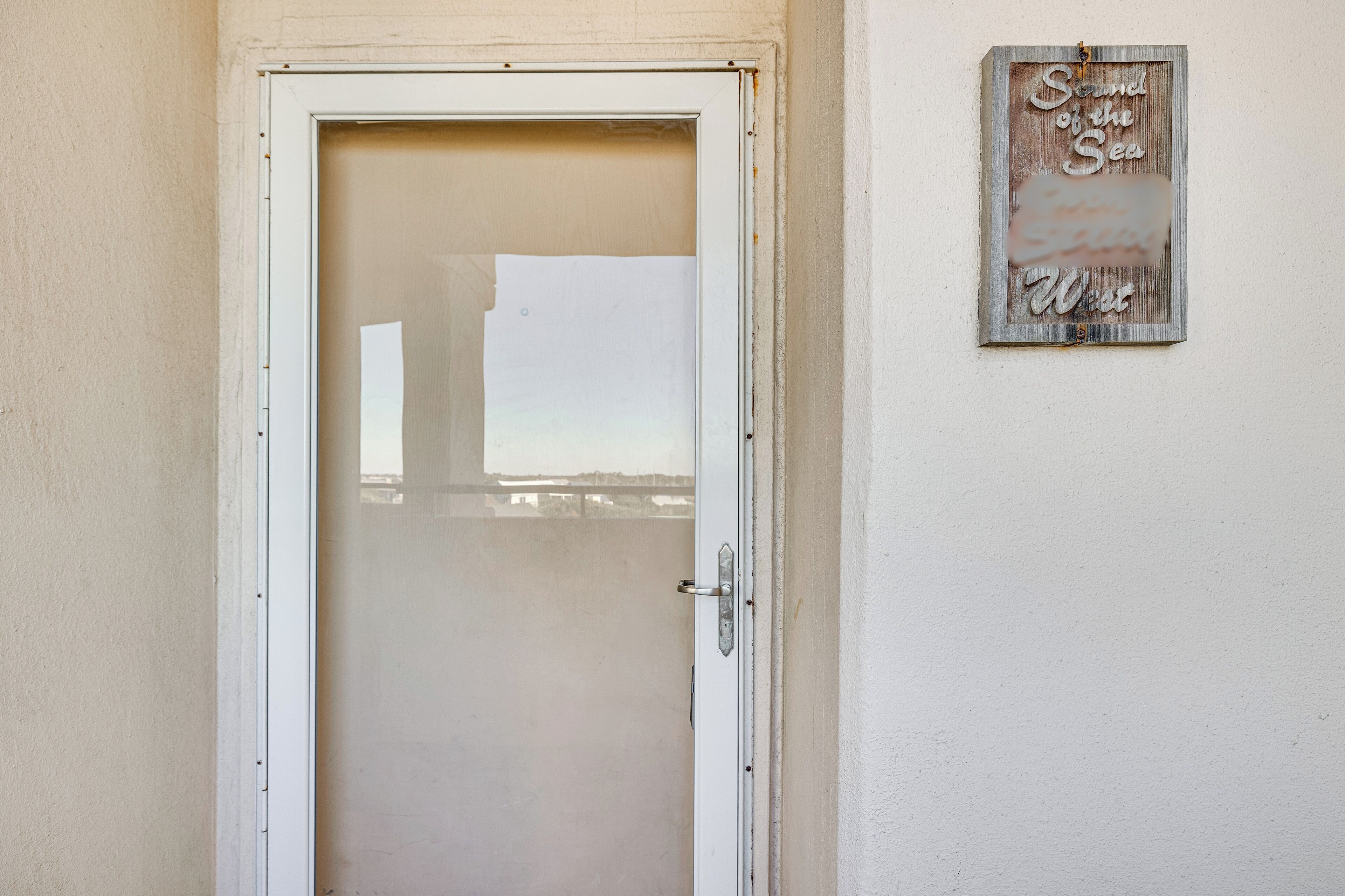 A white door with a frosted glass panel, a metal handle, and a small sign on the wall to the right that reads "Sound of the Sea West".