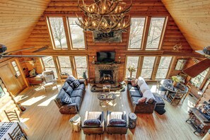 A spacious log cabin interior with a large fireplace, multiple seating areas, and large windows offering views of the outdoors.