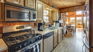 A kitchen with wooden cabinets, stainless steel appliances, and a dining area with a table and chairs. - Reel in Fish, Relax By Fire: Mcminnville Cabin (McMinnville)