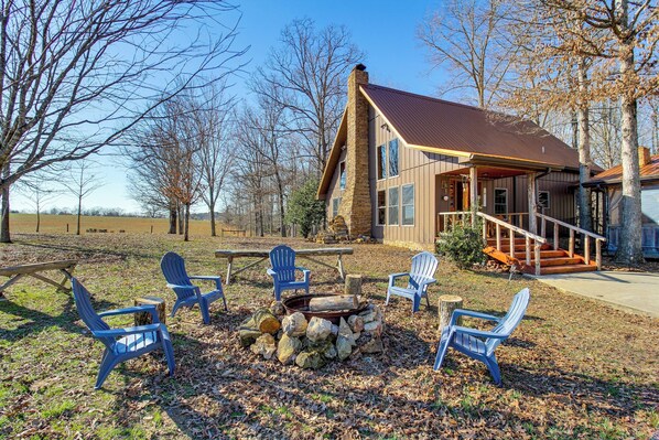 A rustic cabin with a stone chimney, a fire pit, and several Adirondack chairs.