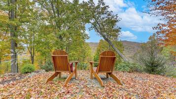 Two wooden Adirondack chairs facing a forested hillside with autumn leaves.