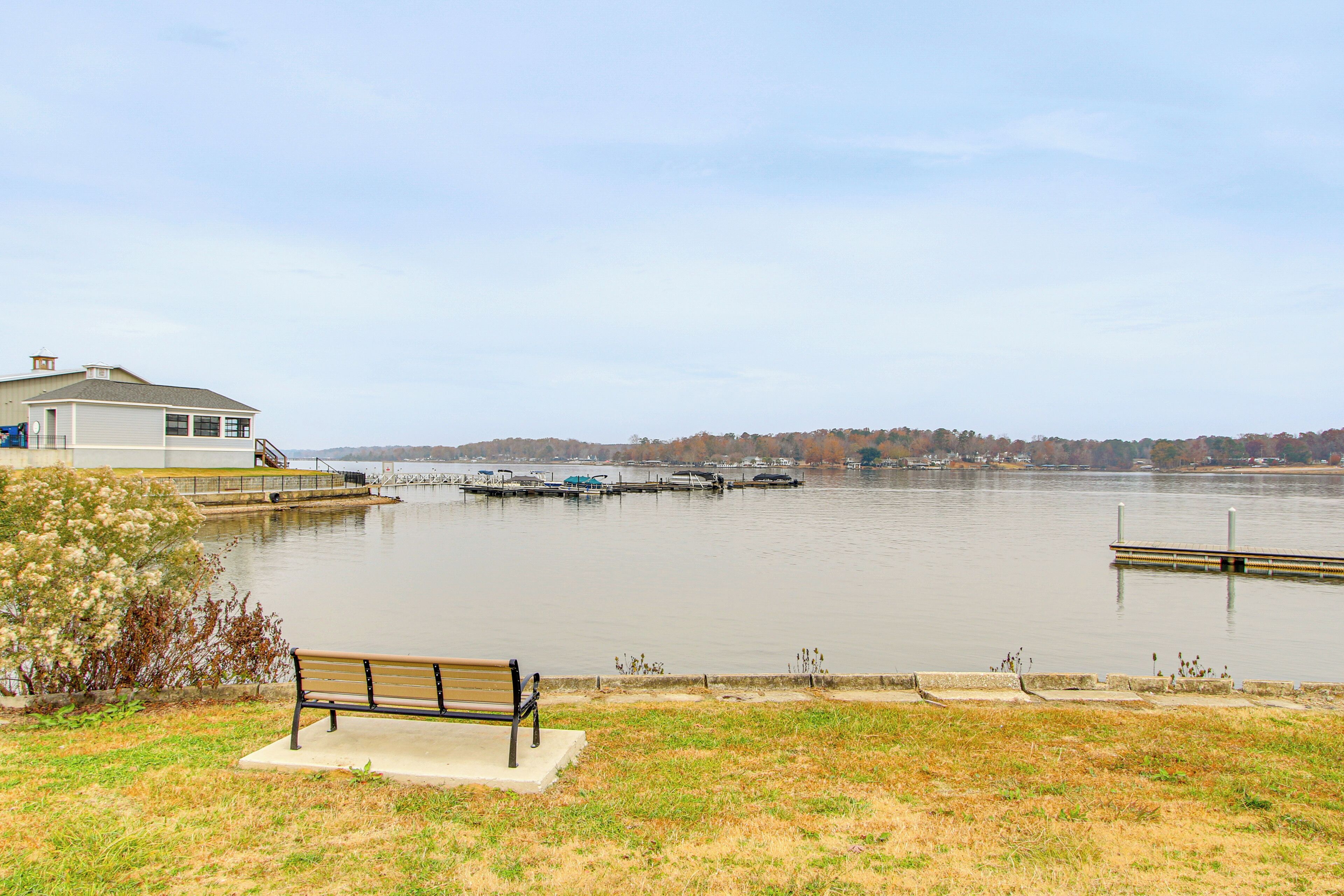 A lakeside view with a bench, dock, and a house near the water.