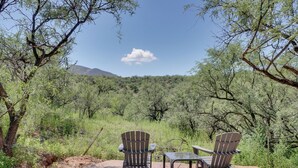 Two Adirondack chairs and a small table set up on a concrete patio surrounded by dense green shrubbery and trees, with a mountain in the background. - Peaceful & Historic Home in Patagonia! (Patagonia)