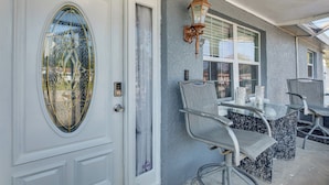 Un porche avec une porte blanche, une fenêtre en verre ornée d’un cadre décoratif, un espace extérieur aménagé avec des chaises et une table en métal, et une lanterne fixée au mur.