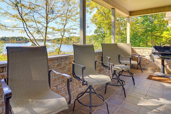 A covered patio with wicker chairs and a grill, overlooking a lake.