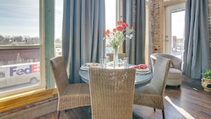 Un comedor con sillas de mimbre, una mesa redonda dispuesta para dos personas, y un jarrón con flores. Se ve una ventana grande con cortinas, y un camión de FedEx en el exterior.