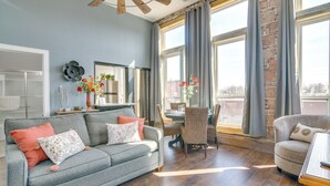 Un salón con un sofá gris, una mesa de comedor con sillas, una mesa de centro de madera y ventanas grandes con cortinas.