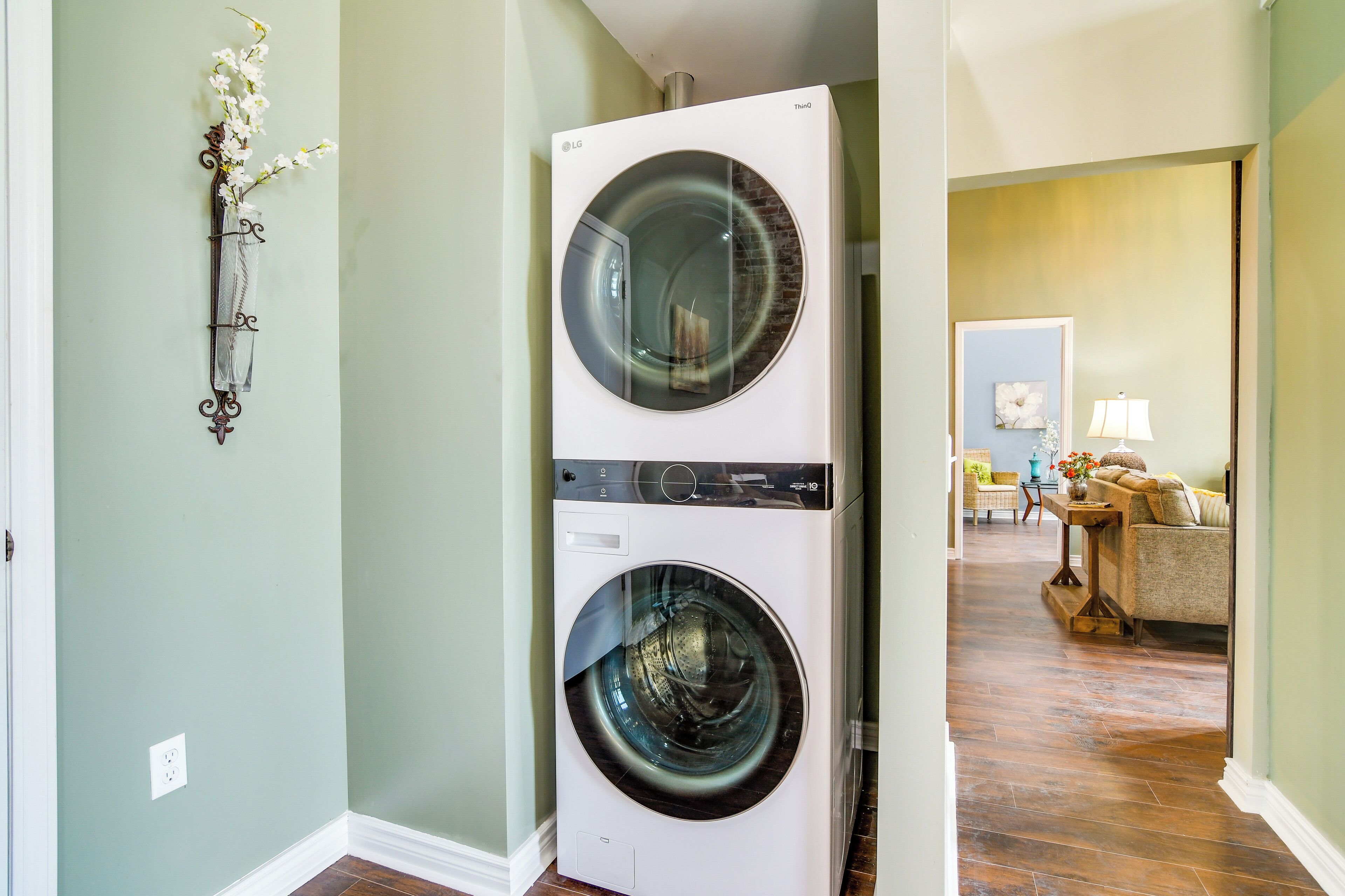 Un lave-linge et un sèche-linge à chargement frontal se trouvent dans une buanderie avec un mur vert, un élément décoratif mural et une vue sur un espace de vie comprenant un canapé et une lampe.
