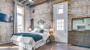 A bedroom with a large bed, a dresser, a TV, and a brick wall. - Dtwn Loft in the Heart of Monett: Pets Welcome! (Monett)