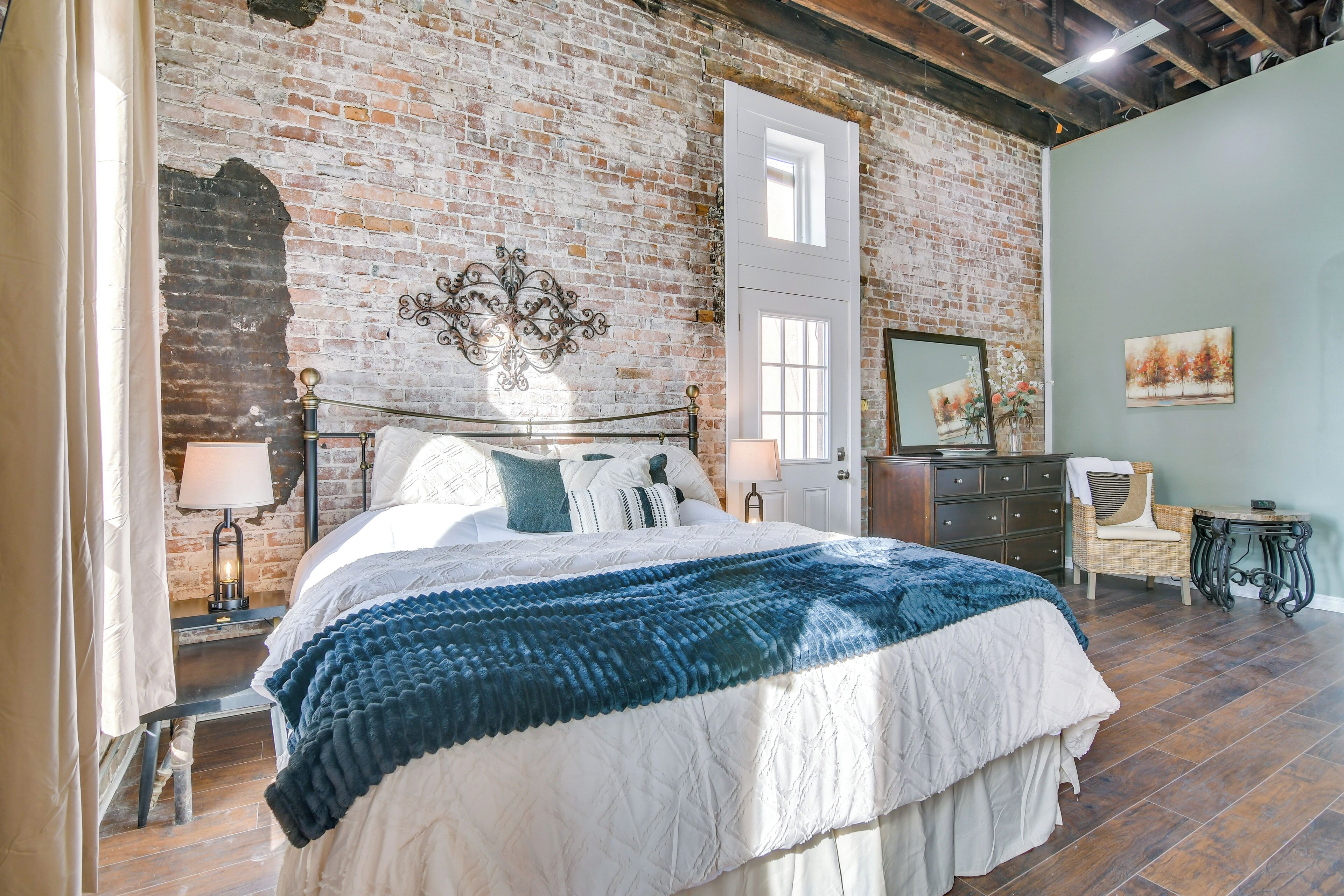 A bedroom with a brick wall, a large bed with a white and blue bedspread, a dresser, a chair, and a painting on the wall.