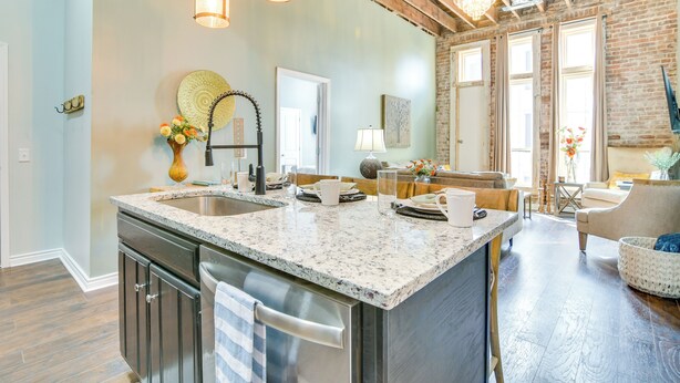 A spacious kitchen with granite countertops, stainless steel appliances, and a large island. There are hanging pendant lights and a brick wall in the background. - Dtwn Loft in the Heart of Monett: Pets Welcome! (Monett)