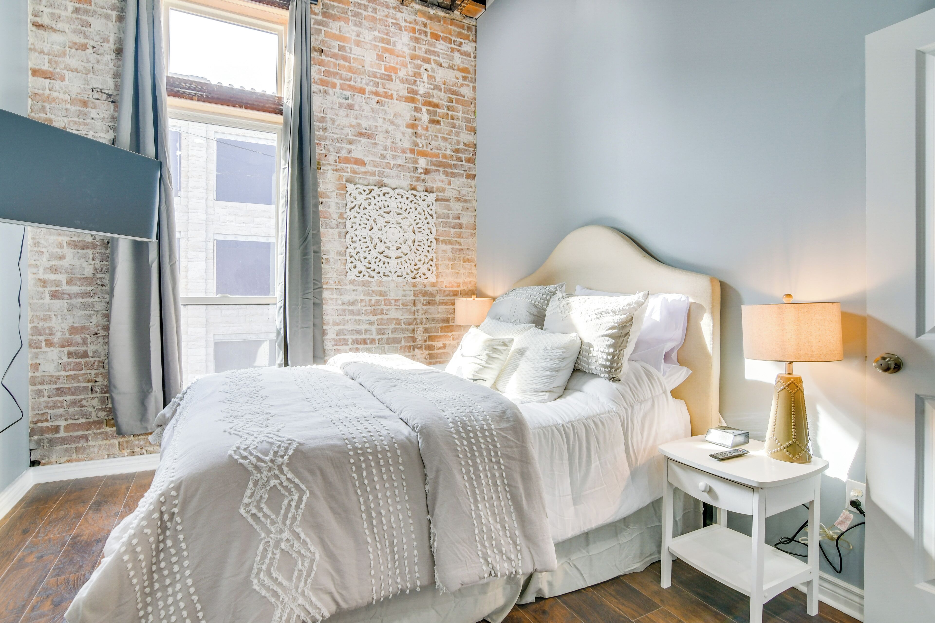 Un dormitorio con pared de ladrillo, cama con ropa de cama blanca, mesita de noche con lámpara y televisor montado en la pared.
