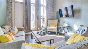 A cozy living room with a brick wall, a large window, a flat-screen TV, and a comfortable seating area with a coffee table. - Dtwn Loft in the Heart of Monett: Pets Welcome! (Monett)