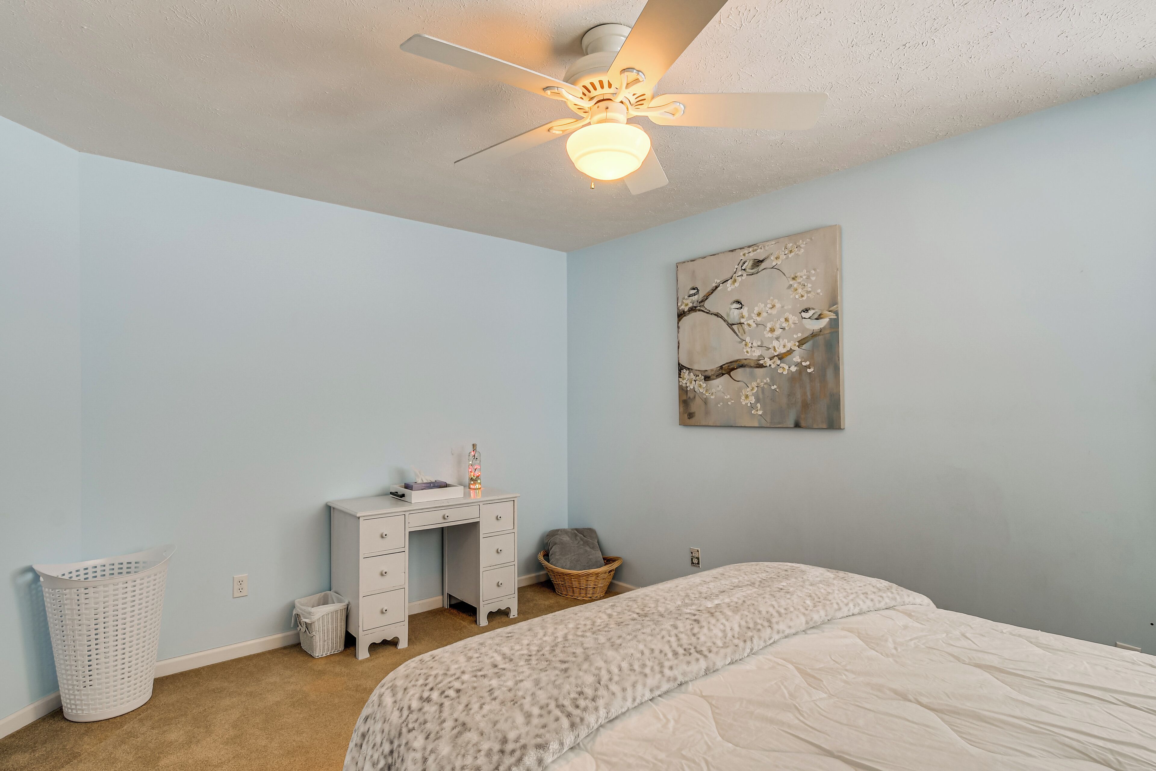 Une chambre à coucher comprenant un lit, un bureau, un ventilateur de plafond, une œuvre d’art murale et un panier à linge.