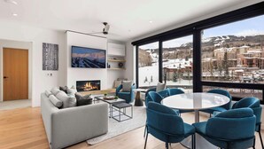 Een moderne woonkamer met een open haard, een groot raam met uitzicht op een besneeuwde berg, en een ronde tafel met stoelen.