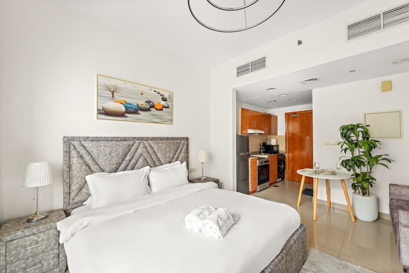 A hotel room with a bed, a bedside table, a lamp, a painting, a small kitchen area, and a potted plant.