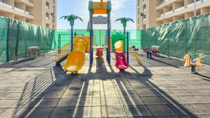 A playground with slides and climbing structures, surrounded by a high fence and palm trees, with a clear sky above. - FIRST CLASS | Studio | Cozy & Chic Retreat (Dubai)