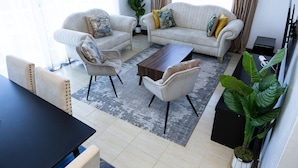A living room with two sofas, a coffee table, and a dining table. There are potted plants and a large window.