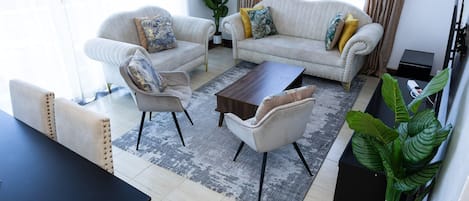 A living room with two sofas, a coffee table, and a dining table. There are potted plants and a large window.