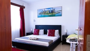 A hotel room with a large bed, a cityscape painting on the wall, a red rug, and a bedside table with a lamp.