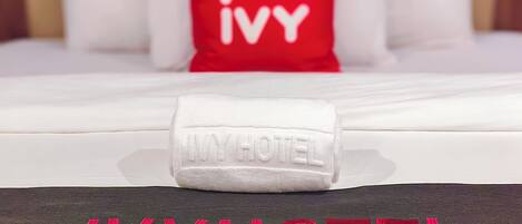 Ein Hotelbett mit weißer Bettdecke, einem roten Kopfkissen mit "Ivy"-Stickerei und einem Handtuch mit der Aufschrift "Ivy Hotel".